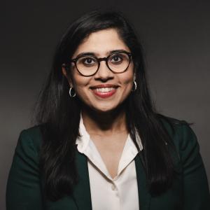 Madhavi Reddi wearing a white shirt, black jacket, and glasses smiling with for her headshot with a black drop