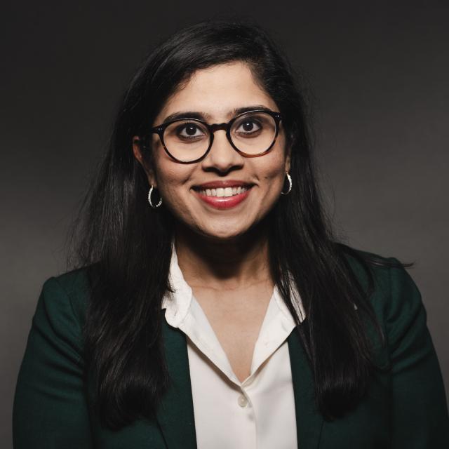 Madhavi Reddi wearing a white shirt, black jacket, and glasses smiling with for her headshot with a black drop